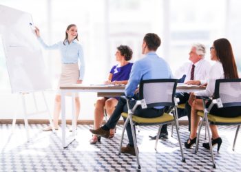 Cheerful young businesswoman pointing at whiteboard and explaining strategy. Confident business coach presenting project to staff. Colleagues listening to presenter. Business meeting concept