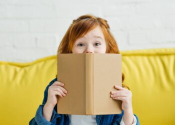 Child sitting on sofa and holding book in front of her face