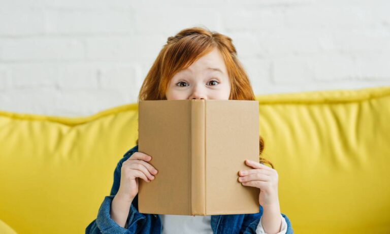 Child sitting on sofa and holding book in front of her face