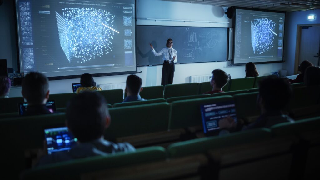 Profesora imparte una clase universitaria sobre inteligencia artificial mostrando gráficos y redes neuronales en pantallas grandes, con estudiantes usando computadoras portátiles en un aula moderna.