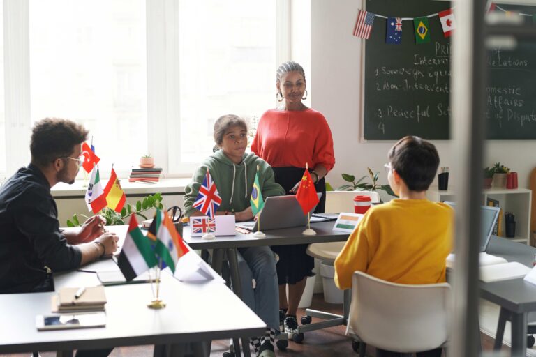 Grupo de estudiantes de diferentes países en un salón de clases moderno con banderas internacionales, participando en una actividad educativa guiada por una profesora.