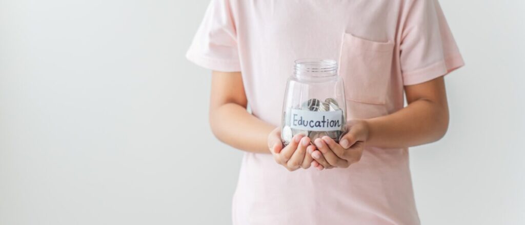 Niño con camiseta rosa sostiene un frasco de vidrio lleno de monedas etiquetado como educación, reflejando la necesidad de recursos frente a recortes educativos.