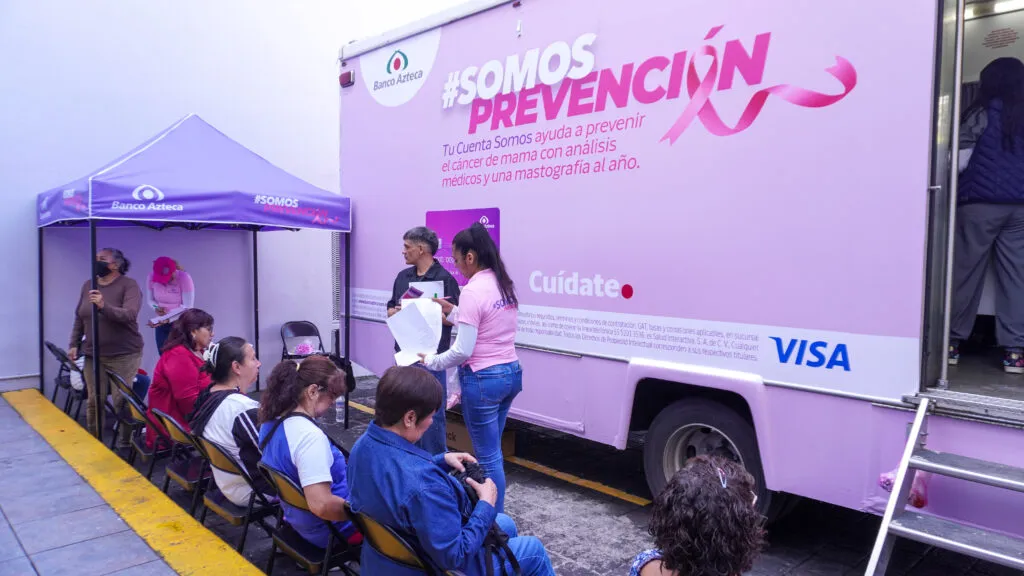 Mujeres esperan frente a una unidad móvil rosa de Banco Azteca, identificada con la campaña SOMOS Prevención.